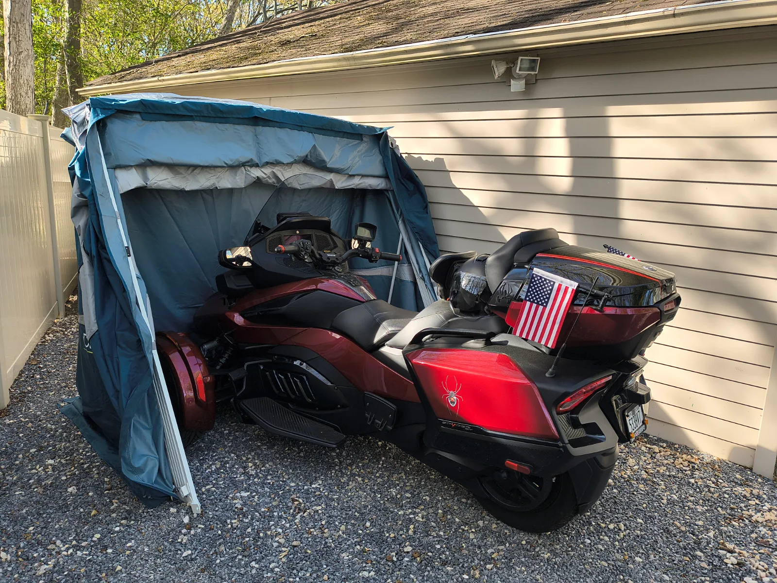 Red Can-Am Spyder parked in an open Deluxe Speedway Shelter on gravel beside a house