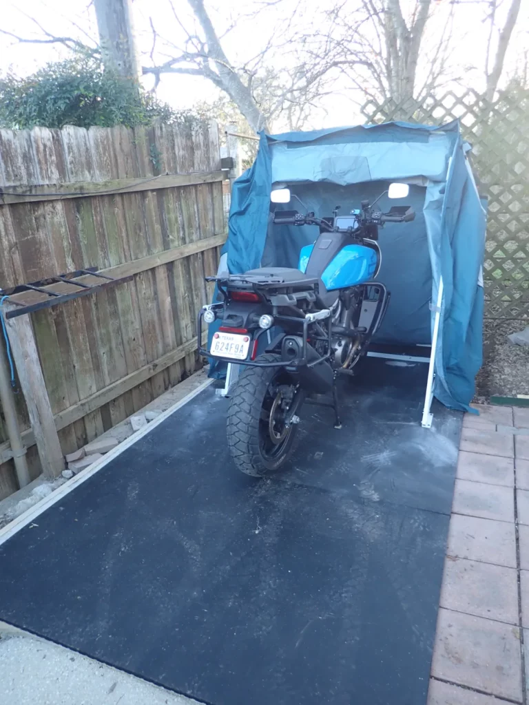 Motorcycle being rolled into an open Speedway Shelter on rubber flooring beside a fence